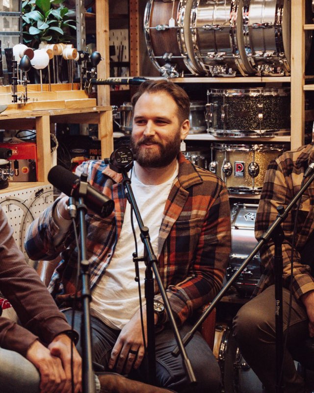 Dan laughing mid-conversation in a drum shop, shelves of snare drums behind him.